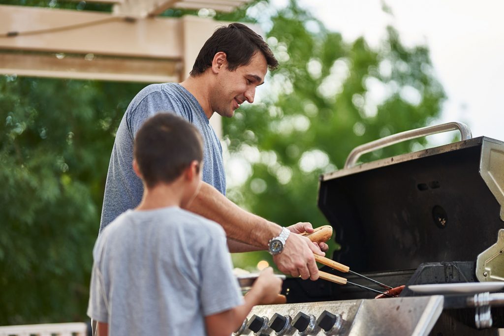 A man grills food using tongs while standing next to a child on a patio, surrounded by lush greenery and wooden pergola beams overhead.