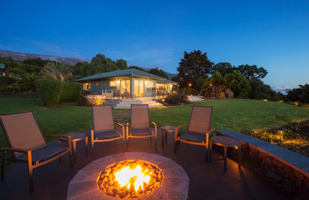 Chairs positioned around a lit firepit sit on a patio; in the background, a house is surrounded by a well-kept lawn, trees, and a dusk sky.