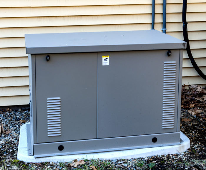 A gray electrical generator sits on a concrete pad. Cables connect it to a control box mounted on yellow siding panels.