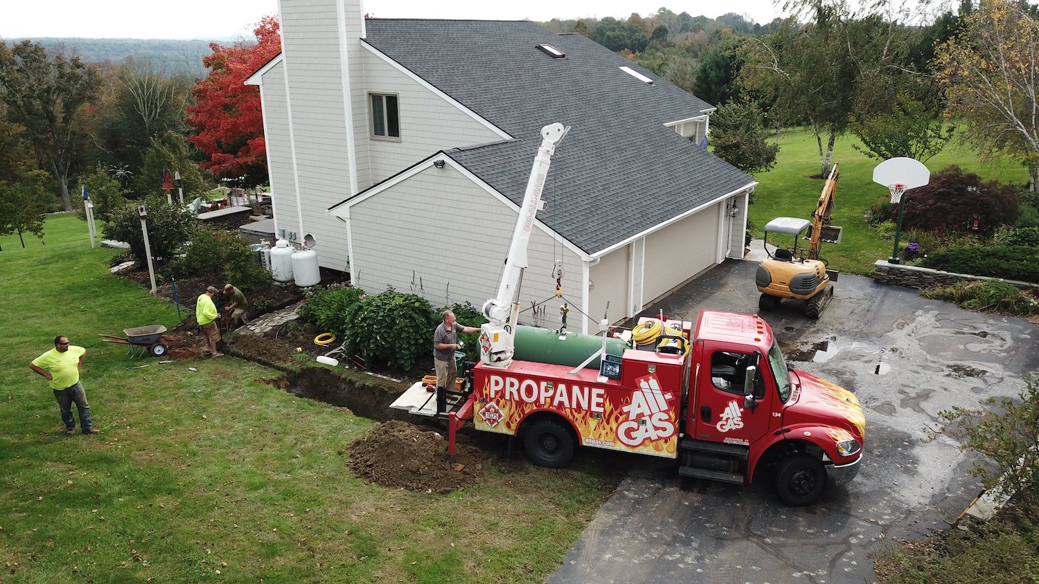 A red propane truck labeled "All Gas" with a crane is delivering a green tank to a residential house. Workers stand nearby.